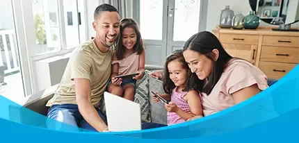 A smiling family of four shares a tablet together on a bright living room couch.